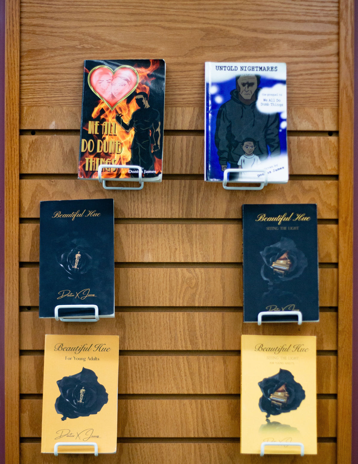 Six books by Dontez X. James displayed on wooden shelves, including Beautiful Hue, Seeing the Light, We All Do Dumb Things, Untold Nightmares, and Young Adult editions.